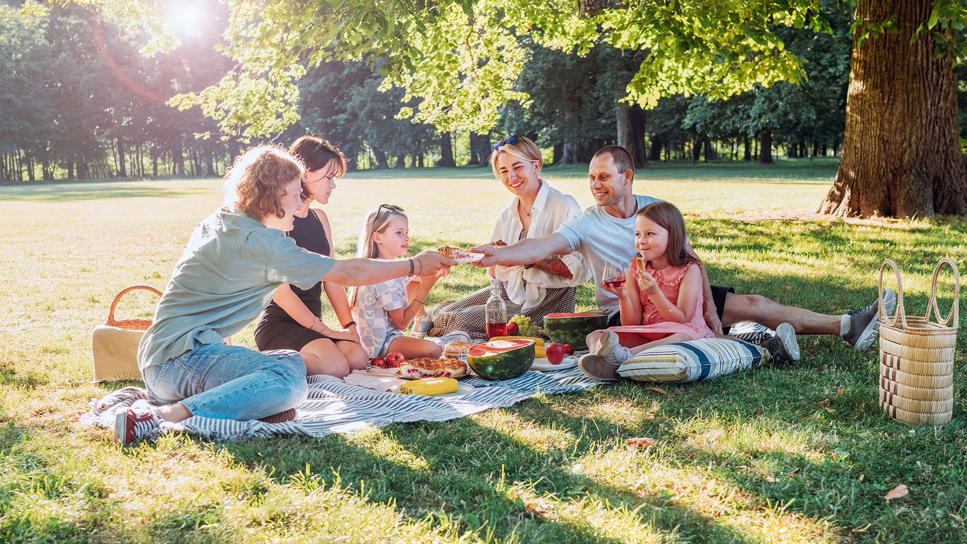 Drei Frauen, ein Mann und zwei Mädchen sitzen auf einer Picknickdecke auf einer Wiese neben einem Baum und essen.