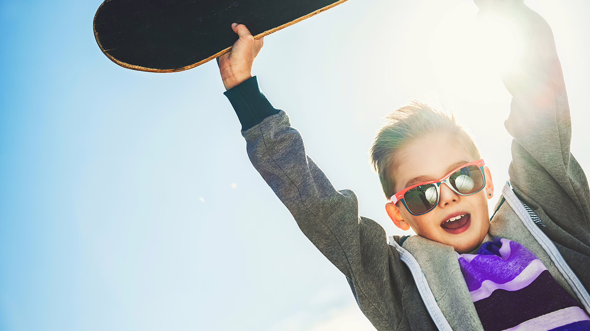 Ein Junge mit einer Sonnenbrille hebt ein Skateboard über seinen Kopf. Im Hintergrund scheint die Sonne.
