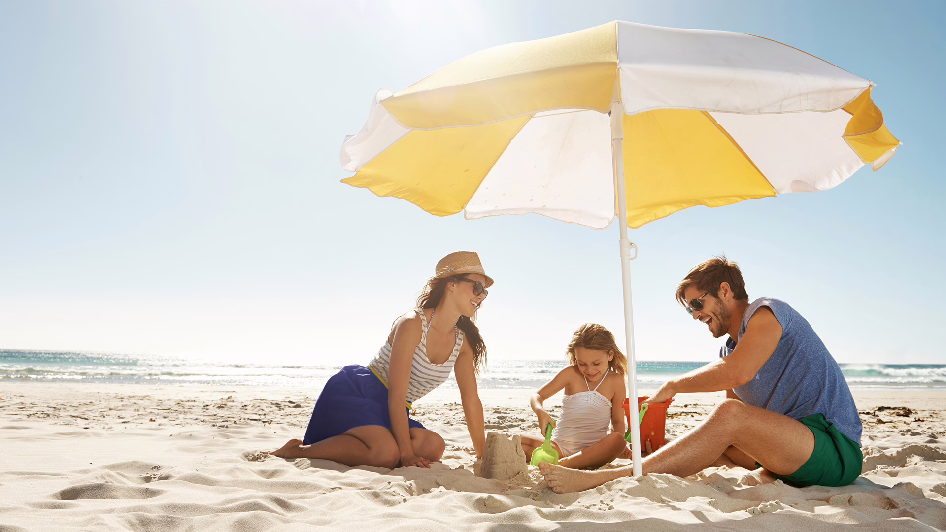 Eine Frau, ein Mann und ein Mädchen sitzen unter einem Sonnenschirm am Strand und bauen eine Sandburg