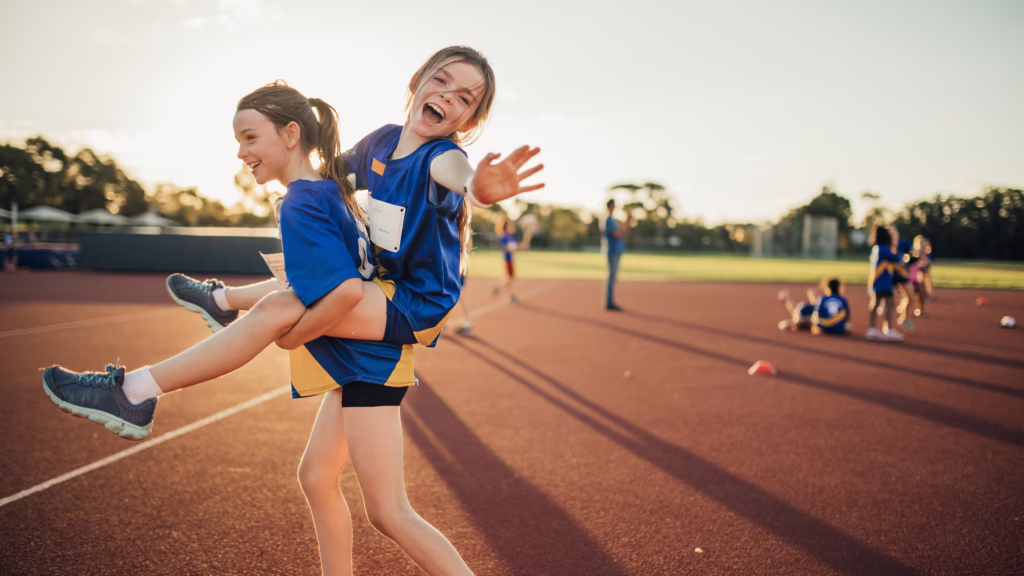 Mädchen trägt anderes Mädchen Huckepack auf einem Sportplatz.