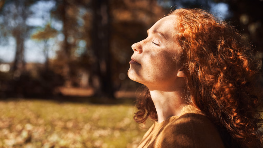 Eine Frau mit roten, lockigen Haaren und geschlossenen Augen genießt ein Sonnenbad im Park.