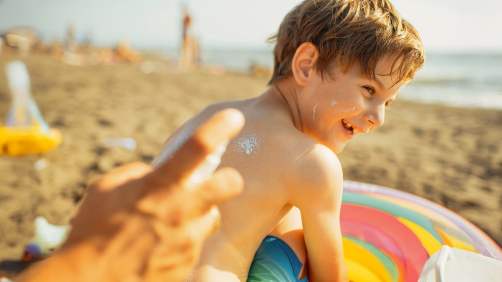 Ein Junge sitzt am Strand und wird am Rücken mit Sonnencreme eingecremt. Von der zweiten Person sind nur die Hände zu sehen.