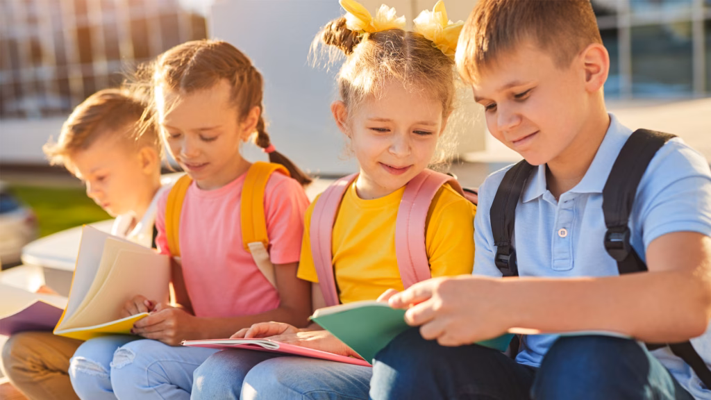 Gruppe jugendlicher Klassenkameraden mit Notizbüchern, die Hausaufgaben machen, während sie in der Nähe des Schulgebäudes in der Sonne sitzen.