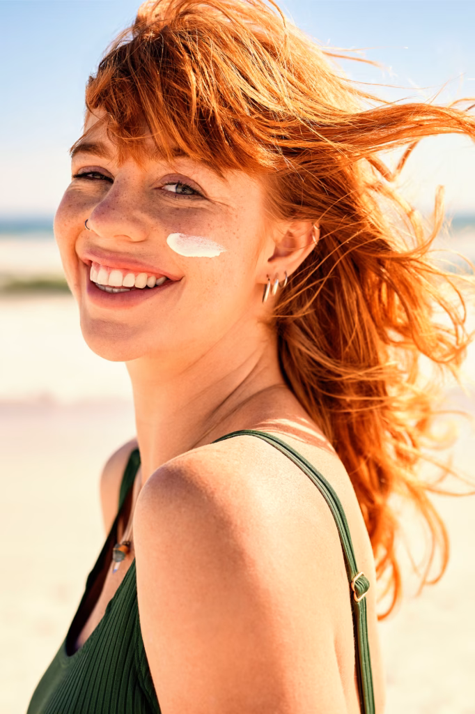 Lächelnde junge Frau, die sich mit Sonnencreme auf der Wange am Strand sonnt.