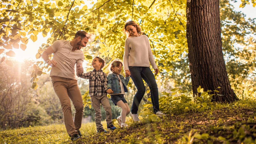 Eine Familie – Vater, Mutter und zwei Kinder – gehen fröhlich in einem Park unter Bäumen spazieren, die Sonne scheint.